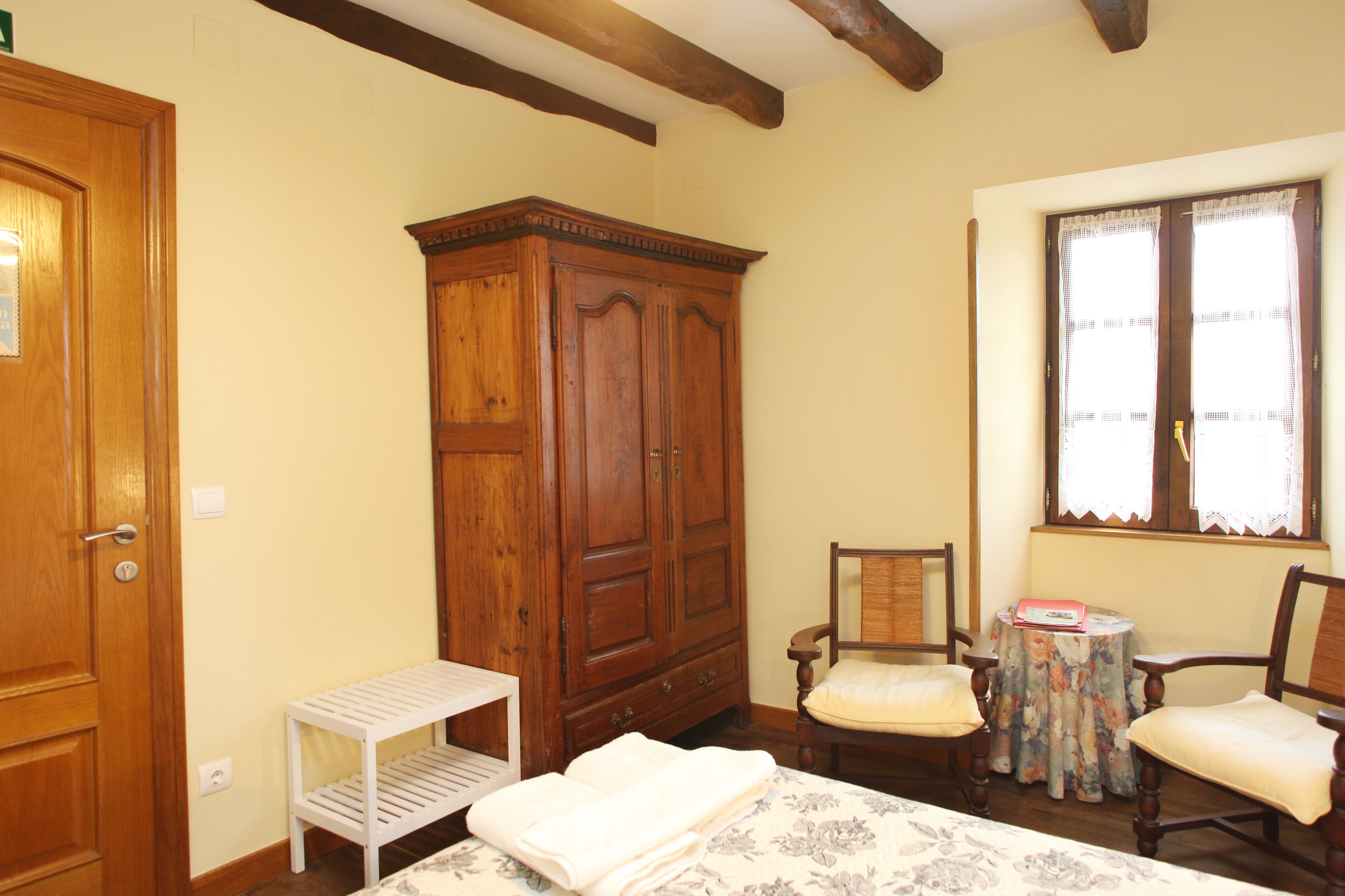 A bedroom with a wooden wardrobe, a bed with a floral bedspread, two chairs, a small table, and a window with lace curtains.
