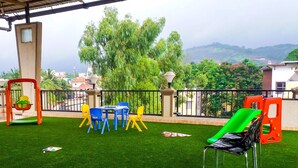An outdoor area with artificial grass, a table with chairs, and a view of greenery and buildings in the background. - Dean Villa (Mawal)