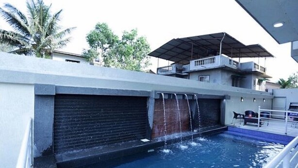 A swimming pool with a waterfall feature, a covered outdoor seating area, and a building with balconies in the background. - Dean Villa (Mawal)
