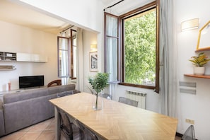 A modern living room with a sofa, a dining table, a TV, and a large window with a view of greenery.