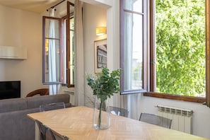 A dining area with a wooden table, clear chairs, a vase with green plants, and large windows with curtains.