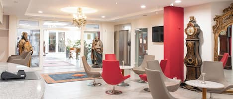 A modern lobby with a reception desk, red and grey chairs, a chandelier, and a large clock.