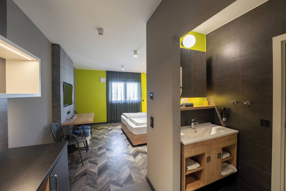 A modern hotel room with a wooden desk, a bed, a large mirror, and a bathroom with a sink and towel rack.