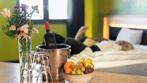 Un letto con una persona sdraiata, una ciotola di frutta, una bottiglia di vino e un vaso di fiori su un tavolo.