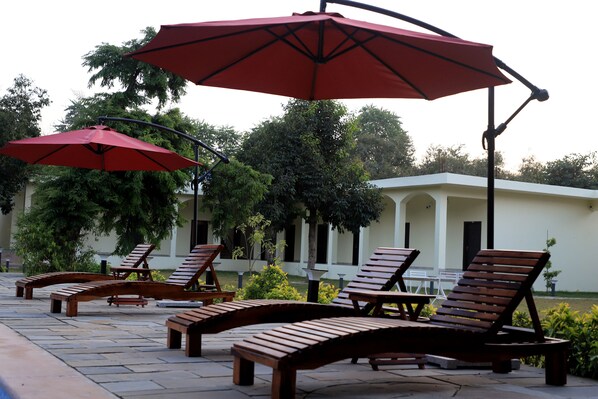 Outdoor lounge chairs and red umbrellas arranged on a patio with a building in the background. - Aroma Resort Pushkar (Pushkar)