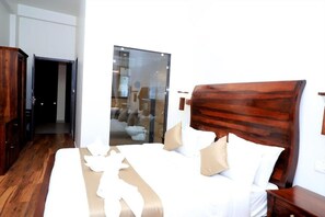 A hotel room with a wooden headboard, a bed with white bedding, a wooden wardrobe, and a glass door leading to a bathroom. - Aroma Resort Pushkar (Pushkar)