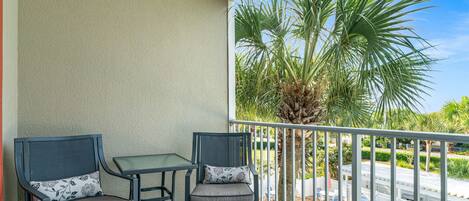 A balcony with two chairs and a small table, overlooking a swimming pool and palm trees.