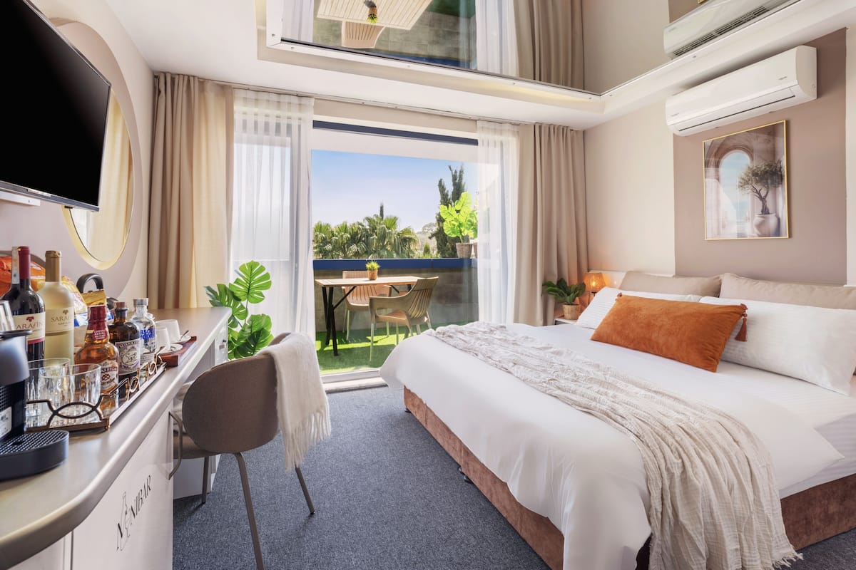 A hotel room with a bed, a desk with a computer, a TV, and a balcony with plants.