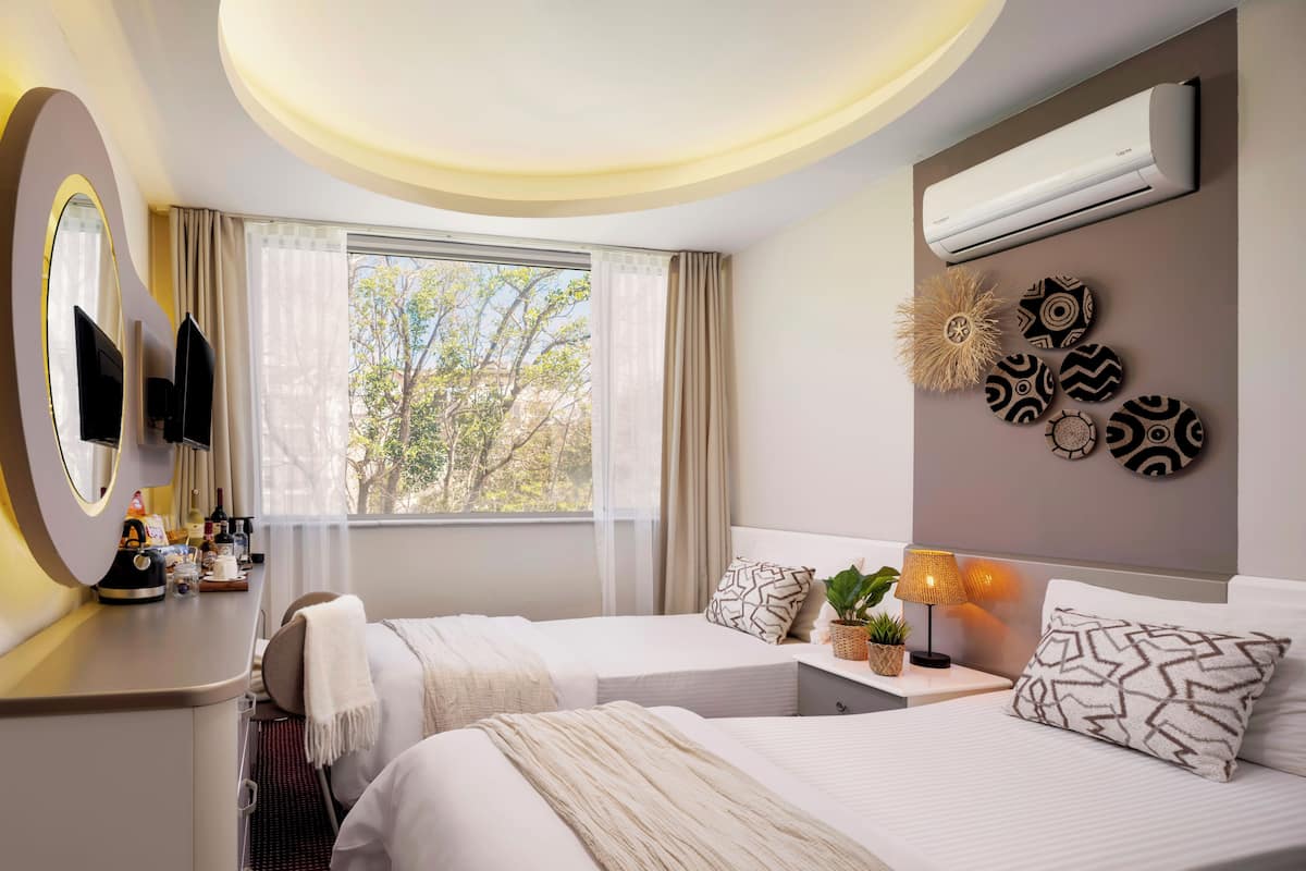 A hotel room with two beds, a TV, an air conditioner, a window with curtains, and a wall with decorative plates.