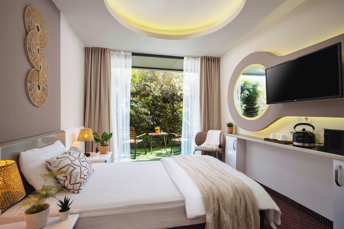 A modern hotel room with a bed, bedside table, lamp, potted plants, and a TV.