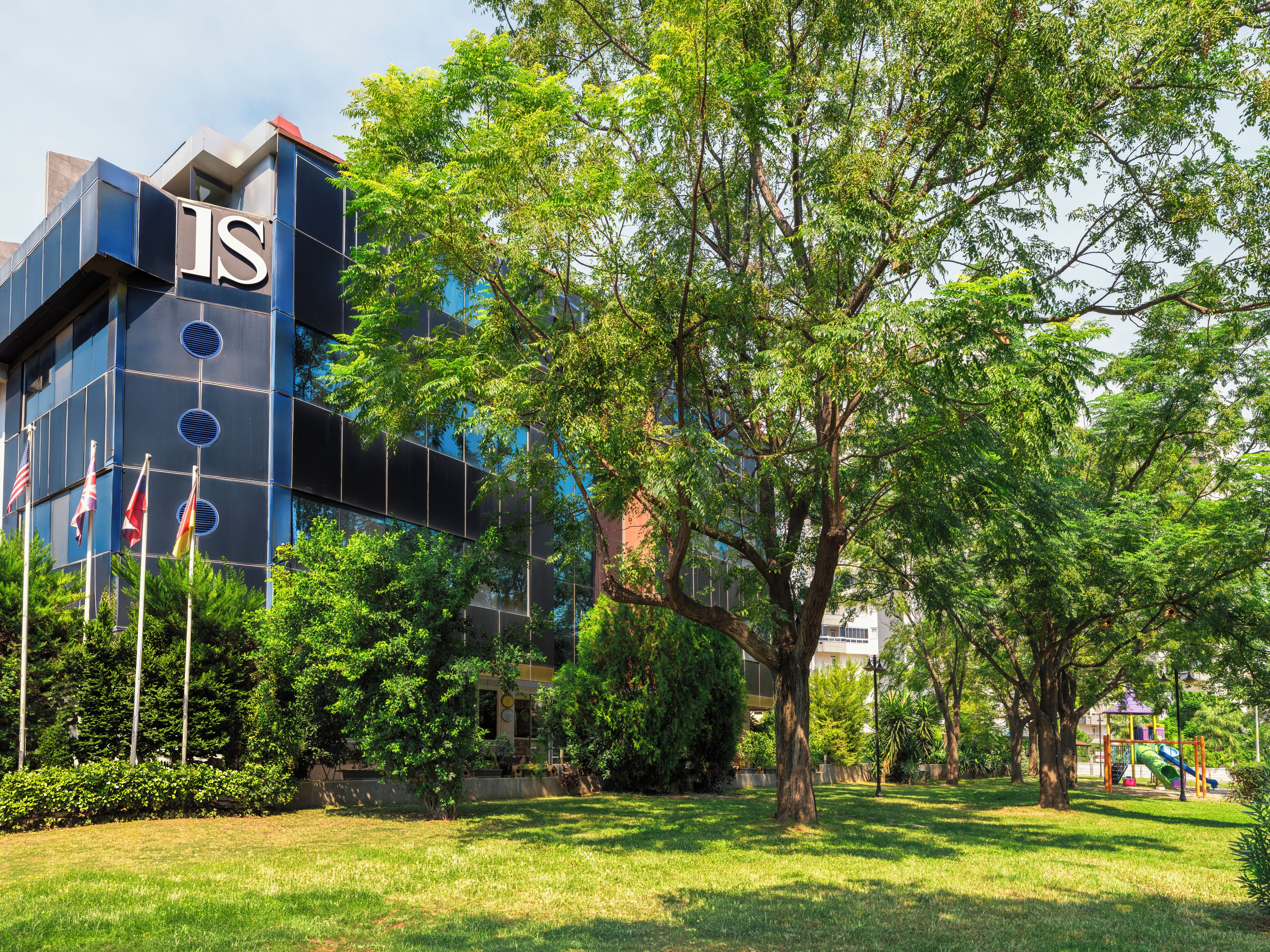 A modern building with a prominent 'IS' logo, surrounded by lush greenery and a playground.