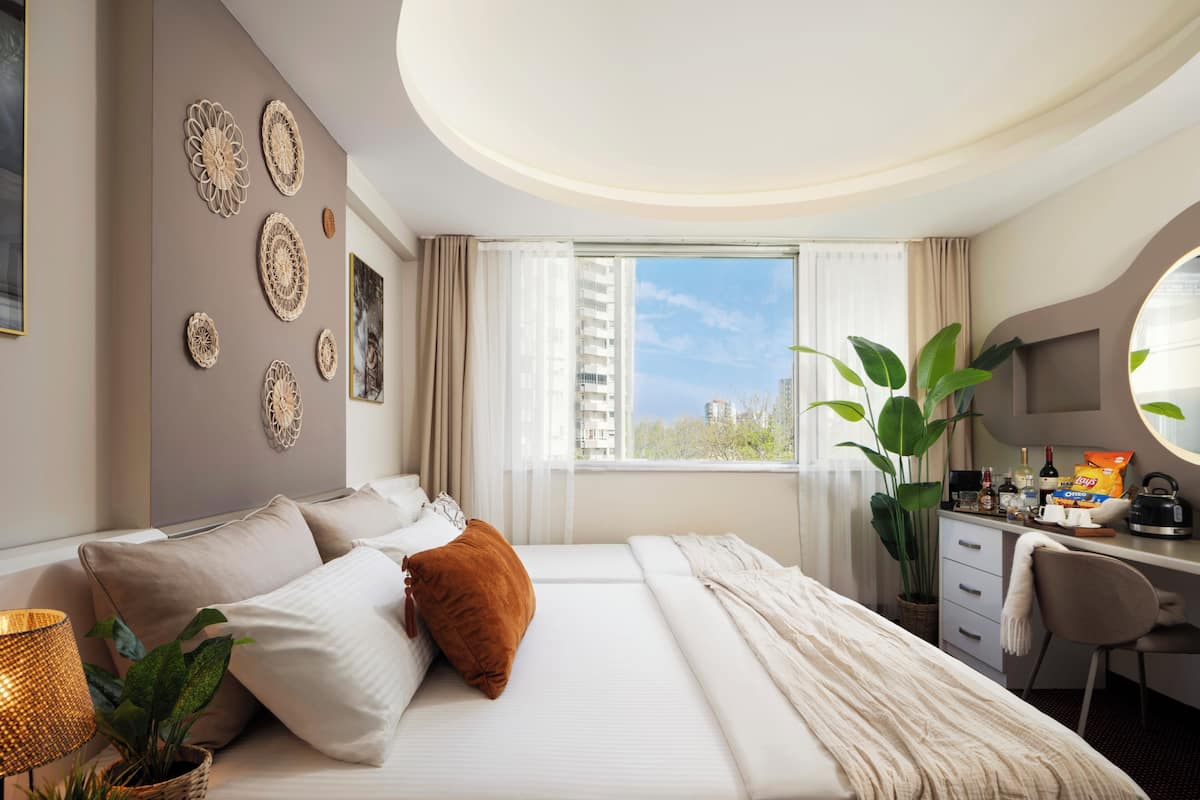 A hotel room with a bed, a desk, a chair, a potted plant, and a view of buildings.