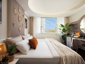 A hotel room with a bed, a desk, a chair, a potted plant, and a view of buildings. - IS Hotel (Antalya)