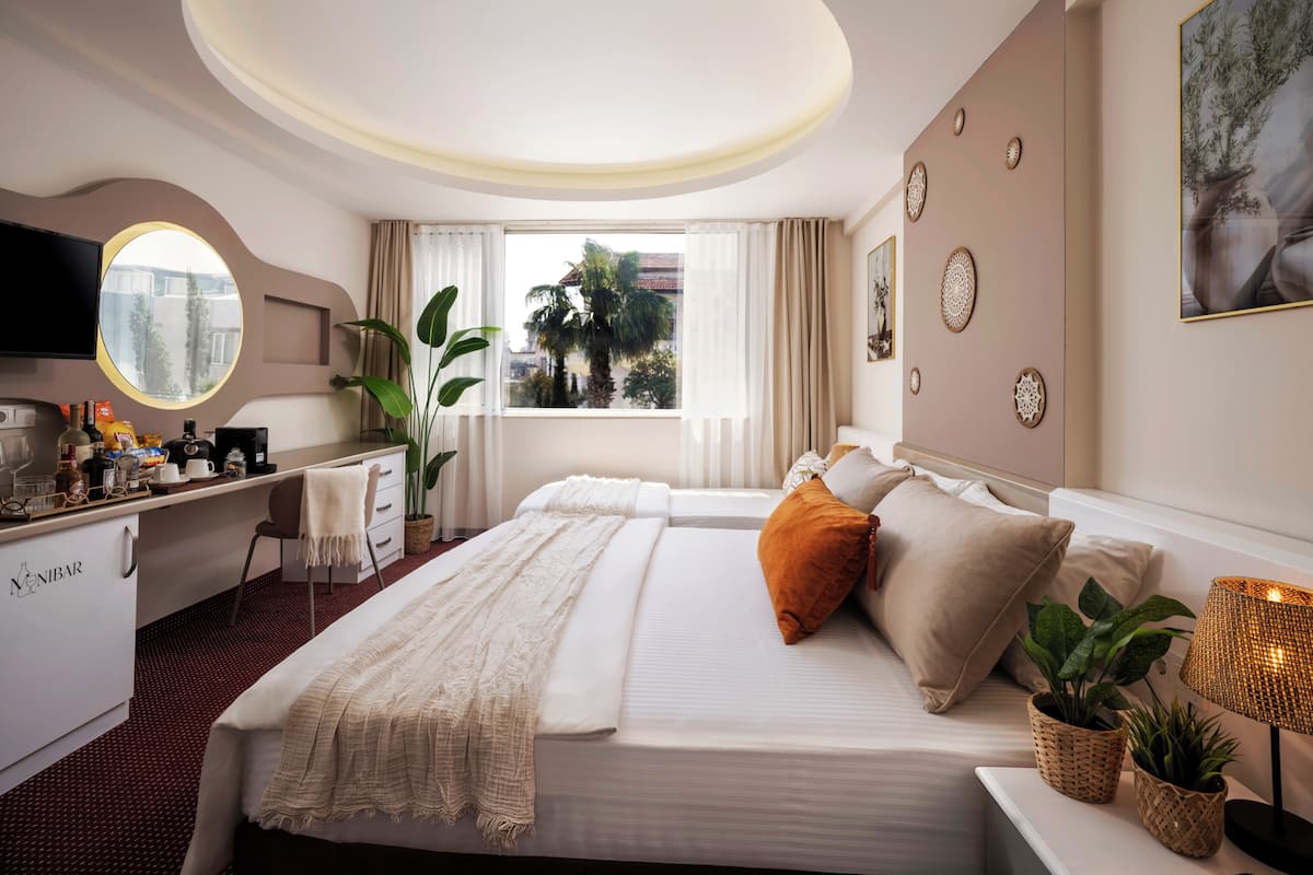 A hotel room with a bed, bedside table, lamp, potted plants, a desk with a computer, and a window with curtains.