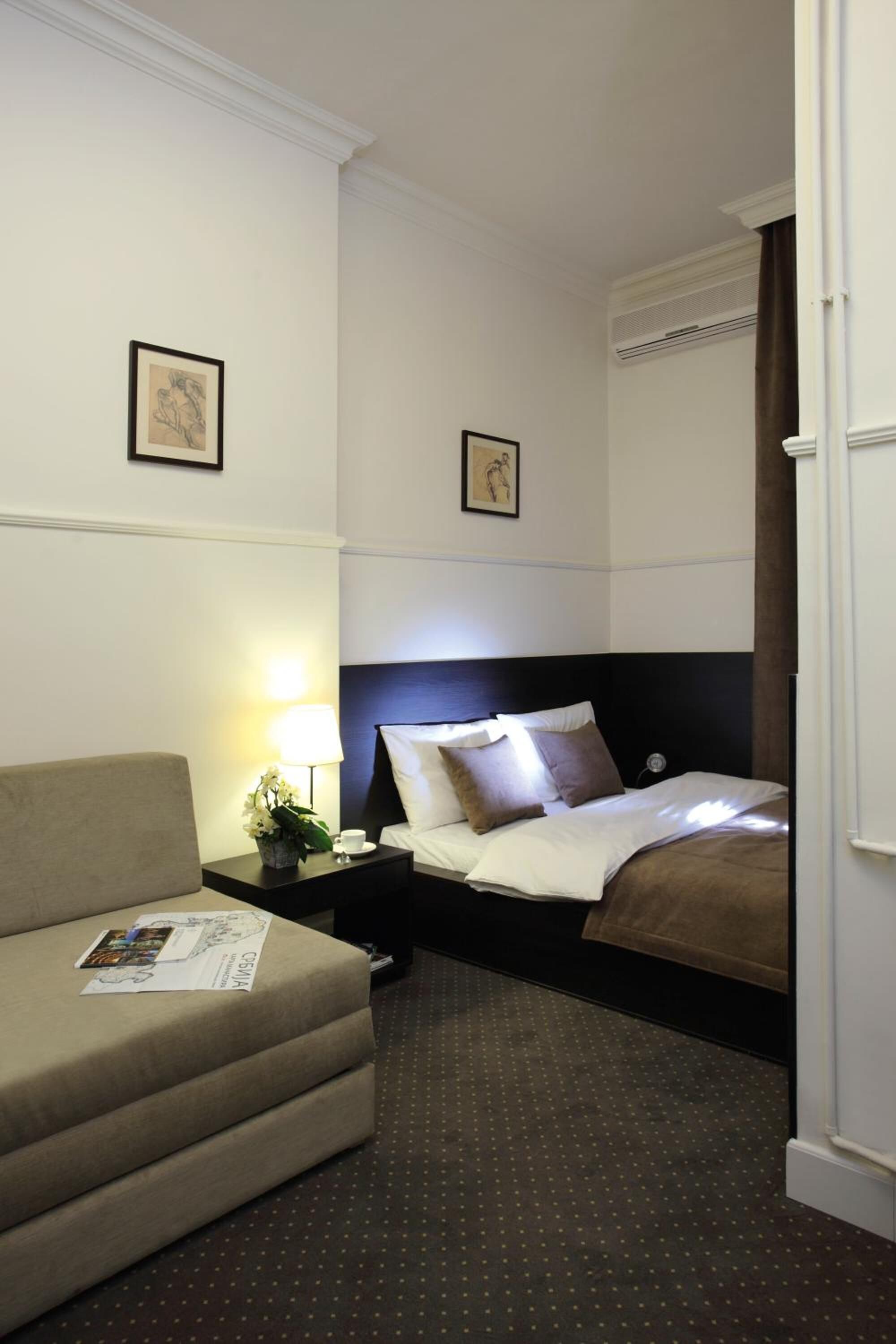 A hotel room with a bed, a sofa, a nightstand with a lamp, and two framed pictures on the wall.