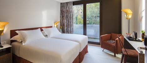 Habitación de hotel con dos camas, un escritorio con silla, una lámpara y vistas al exterior.