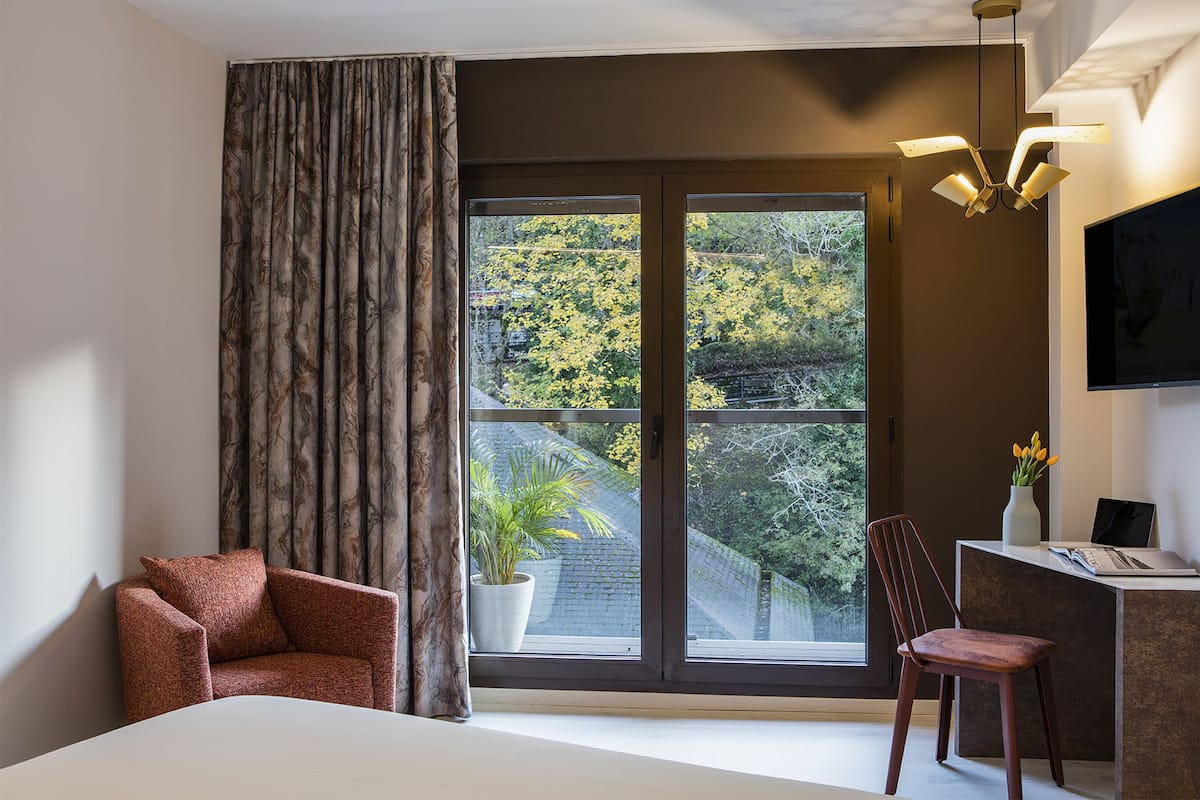 A modern hotel room with a large window, a desk with a laptop, a chair, and a bed.