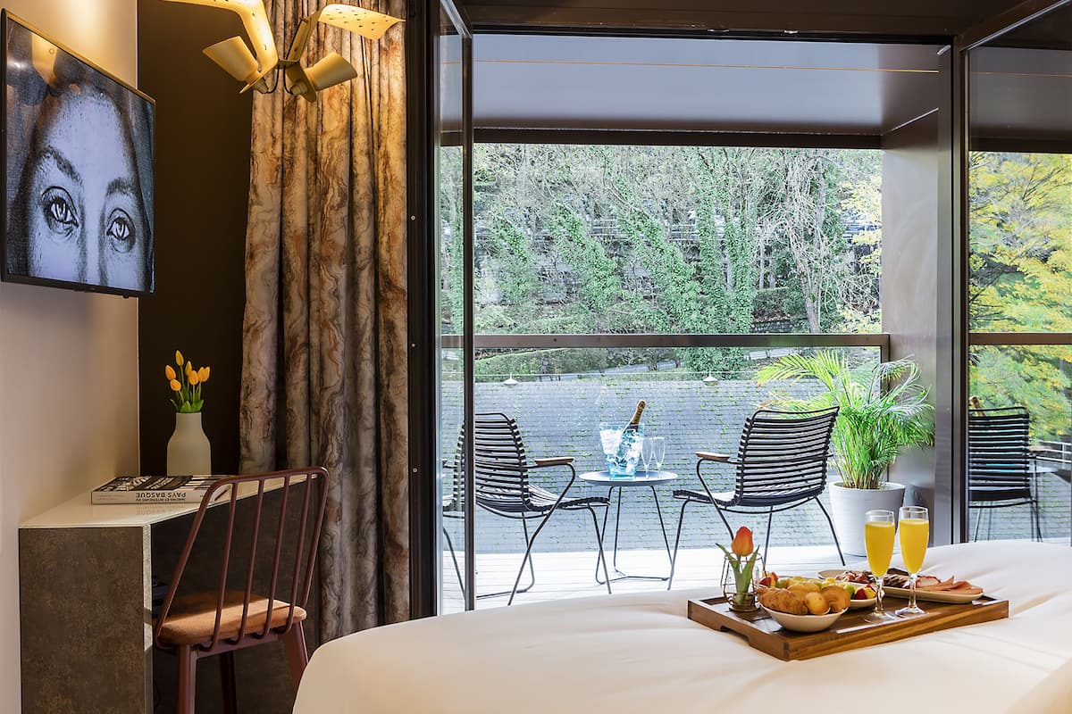 A hotel room with a large bed, a dining area with chairs and a table, a TV, and a view of trees through large windows.