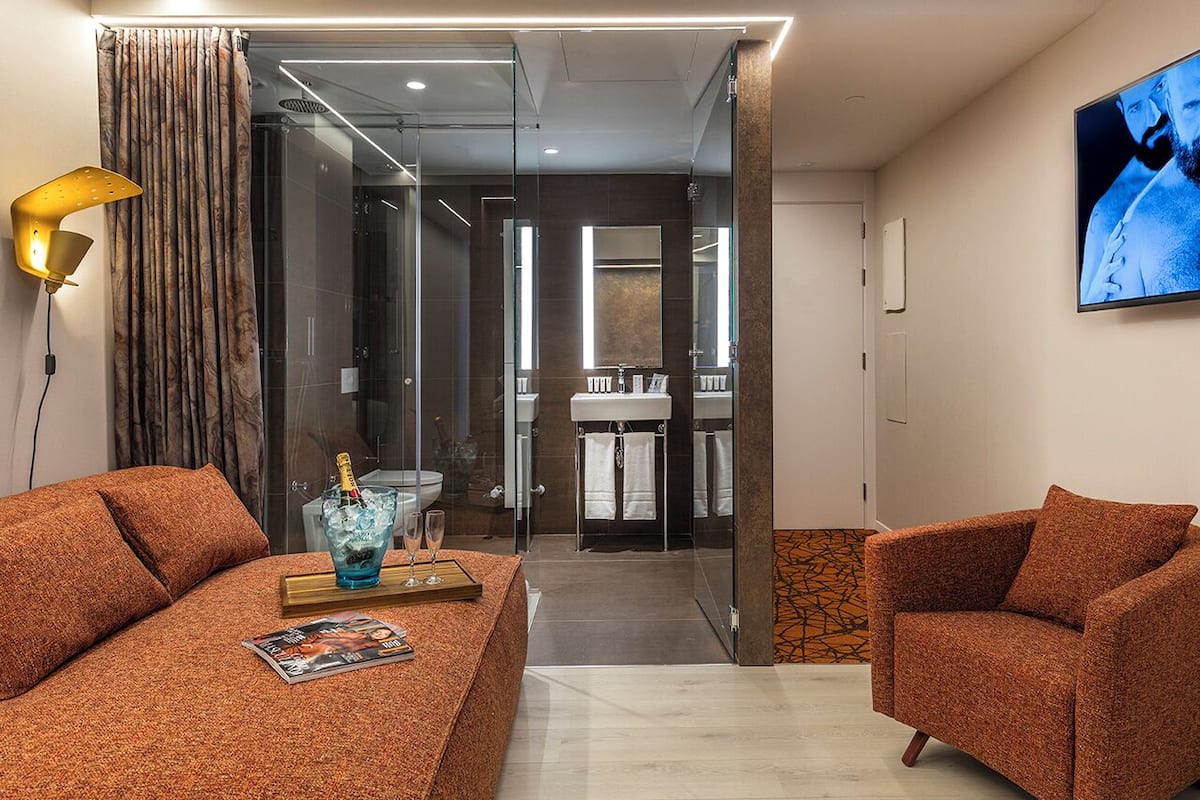 A modern hotel room with a sofa, armchair, and a glass-enclosed shower area.