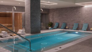 Una piscina interior moderna con una barrera de vidrio, un área de spa con una silla de masaje y una zona de estar con toallas a rayas.
