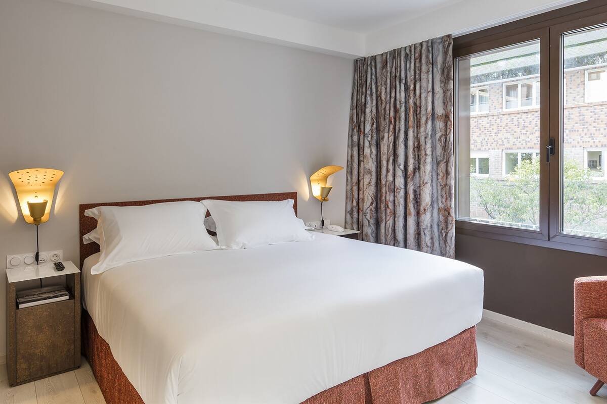 A hotel room with a large bed, two bedside lamps, a nightstand, a chair, and a window with curtains.