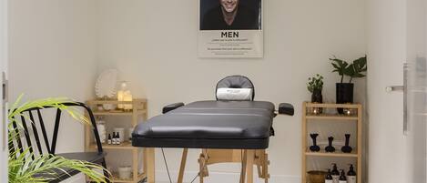 Um quarto de spa com mesa de massagem, uma cadeira, prateleira com frascos e um pôster na parede.