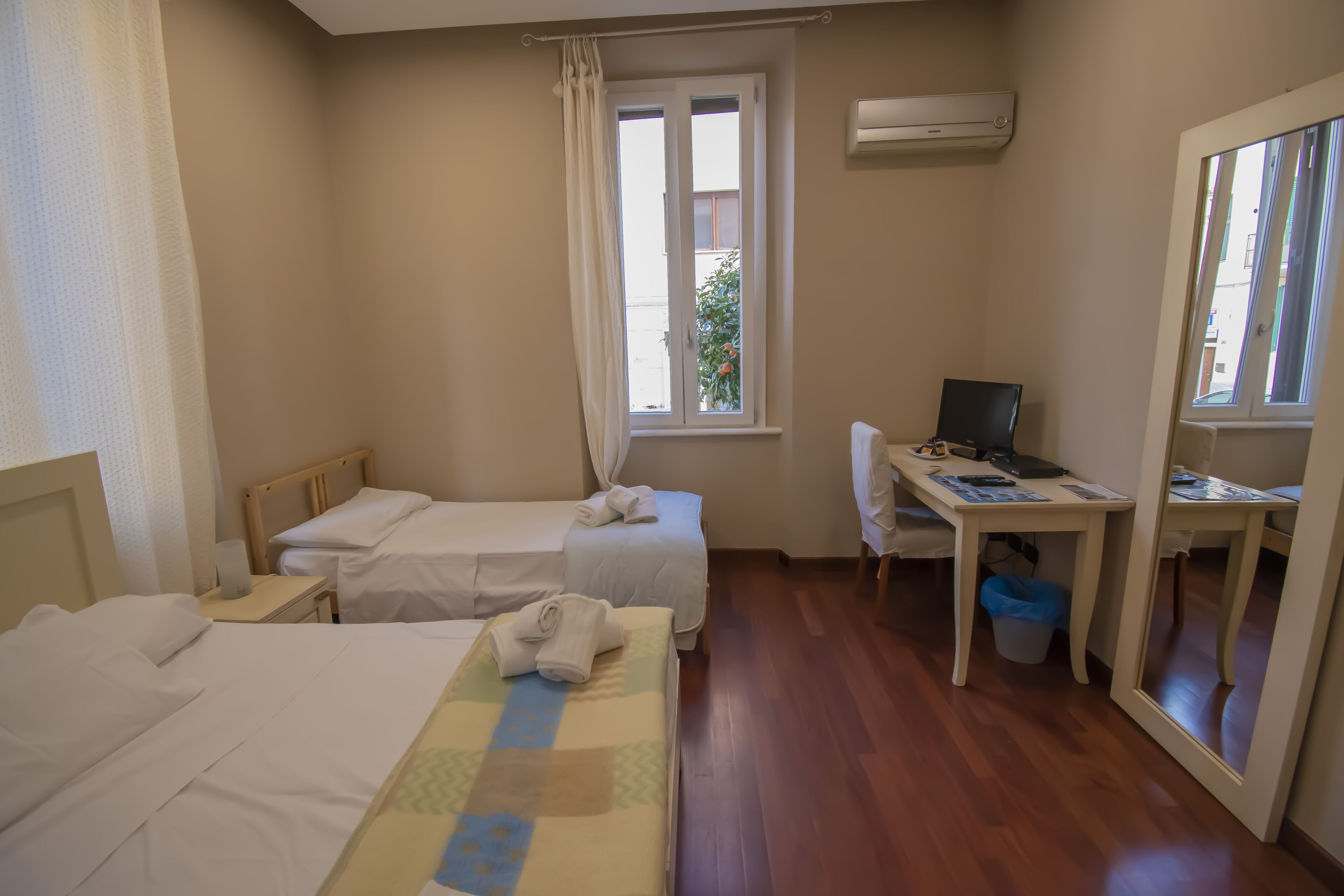A hotel room with two beds, a desk with a computer, a window with curtains, and an air conditioning unit.