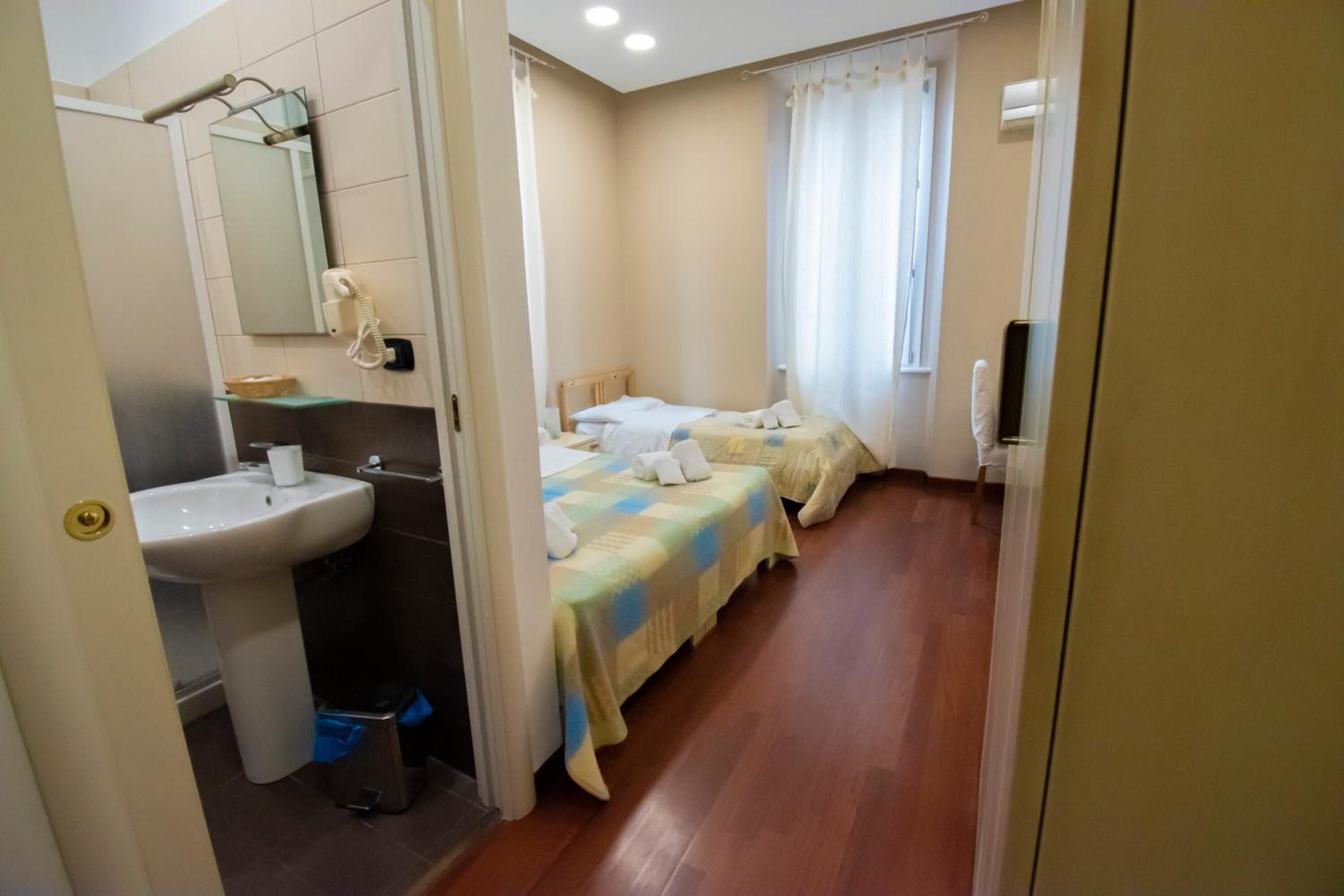 A hotel room with a bathroom, two beds, a bedside table, a mirror, and a window with curtains.