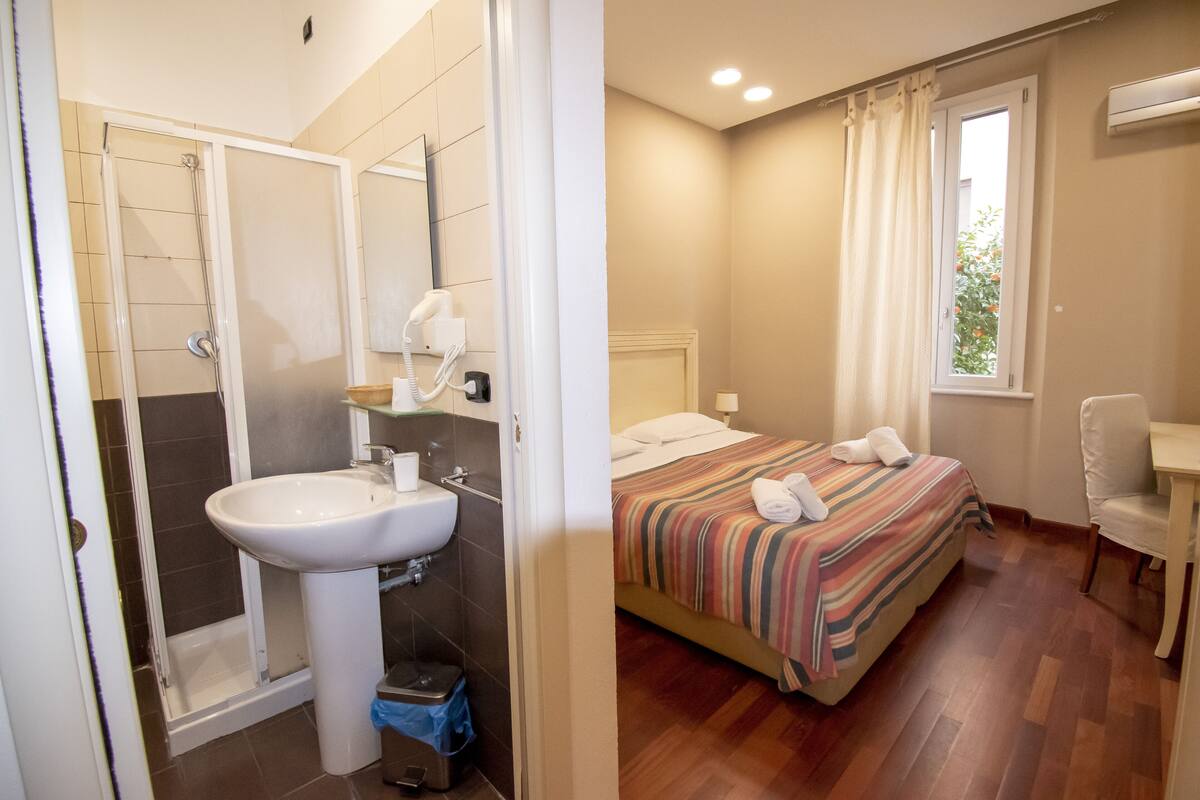 A bathroom with a shower, sink, and toilet. The bedroom has a bed, window, and chair.
