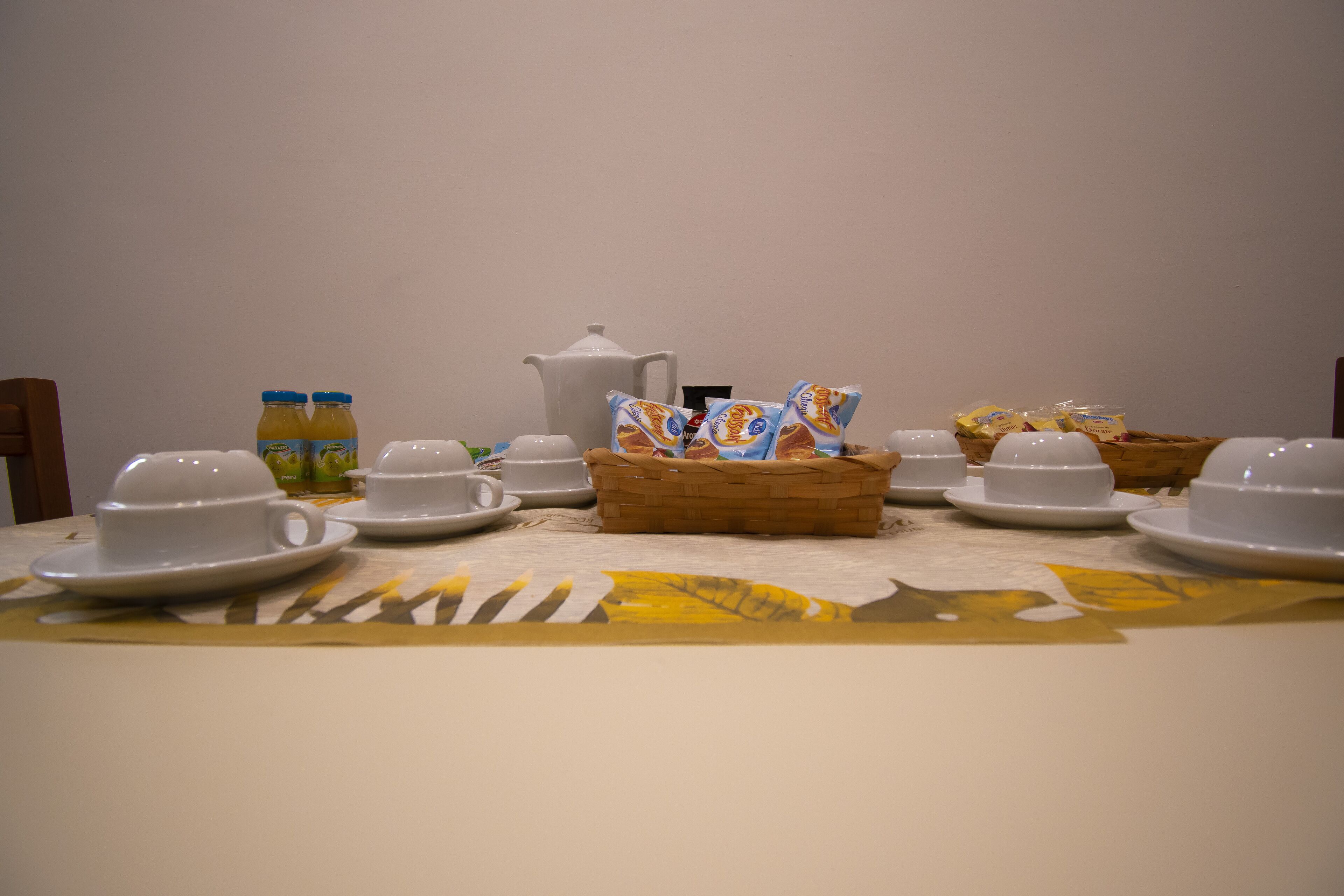 A breakfast setup with white cups, saucers, a teapot, juice bottles, and a basket of pastries on a table with a floral tablecloth.
