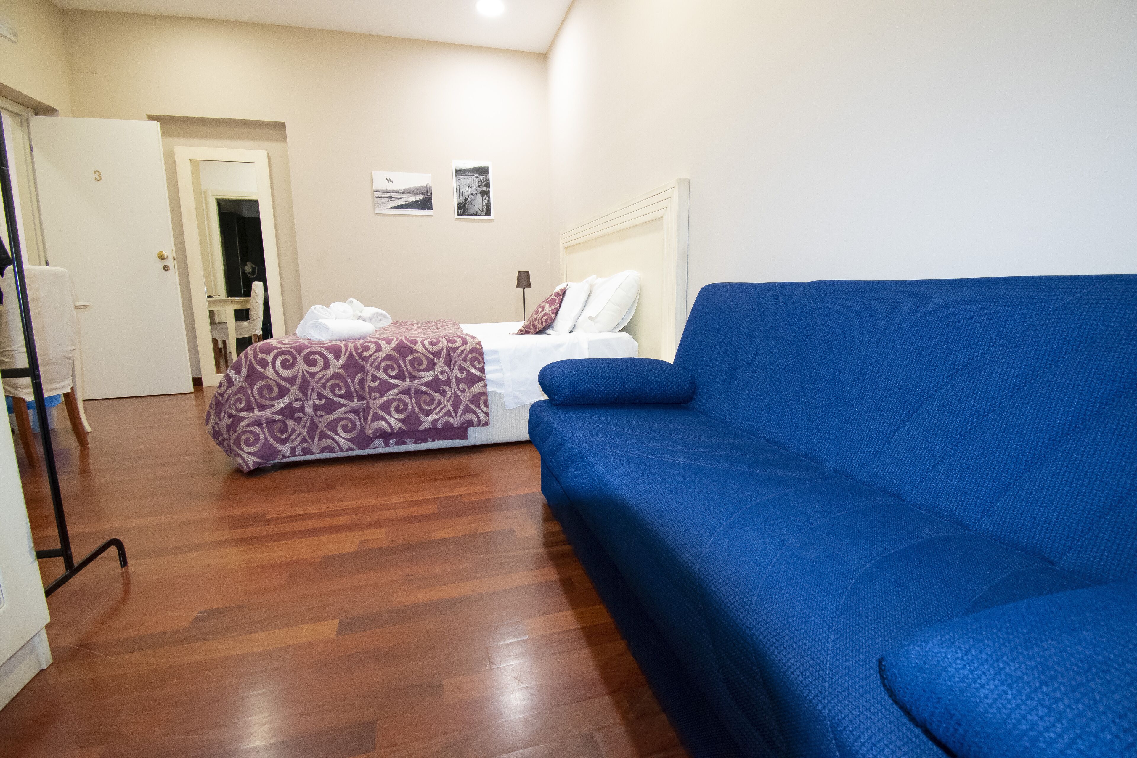 A hotel room with a bed, a blue sofa, wooden flooring, a white door, and a mirror.