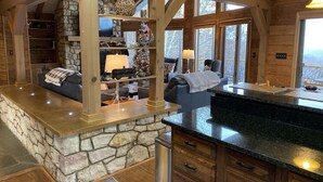 L’intérieur chaleureux d’un chalet avec une cheminée en pierre, des poutres en bois, un comptoir en pierre et une grande fenêtre donnant sur le paysage extérieur.