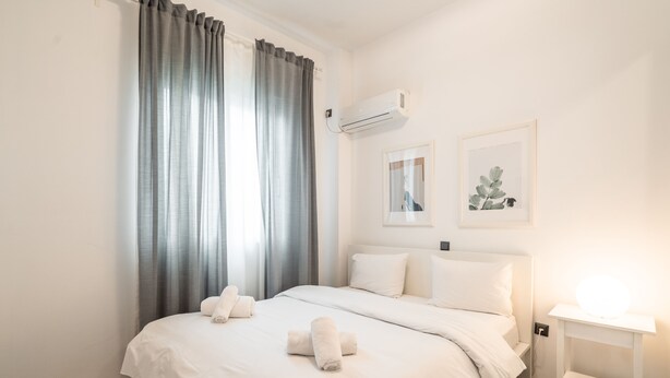 A modern hotel room with a large bed, white bedding, a bedside table with a lamp, and two framed pictures on the wall.