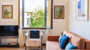 Una sala de estar con televisor, sofá, sillón y ventana con vista a edificios y árboles.