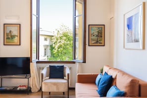 Un salon comprenant un téléviseur, un canapé, un fauteuil et une fenêtre donnant sur des immeubles et des arbres.