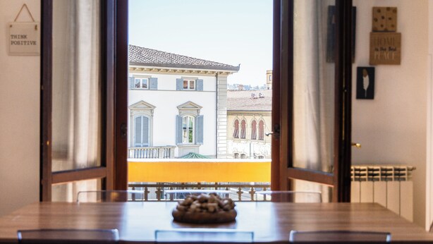 Vista a través de una ventana que muestra una mesa y sillas, un frutero y un edificio de arquitectura clásica.