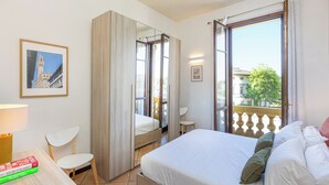 Habitación de hotel con cama, una silla, una mesa con libros, un espejo y un balcón con vistas.