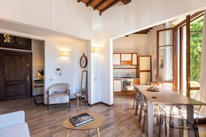 Ampla sala de estar com uma mesa de jantar de madeira, um sofá e uma pequena mesa redonda com um livro. Cozinha ao fundo com eletrodomésticos modernos e uma grande janela.