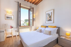 A bedroom with a bed, bedside table, lamp, window, desk, chair, and framed pictures on the walls.