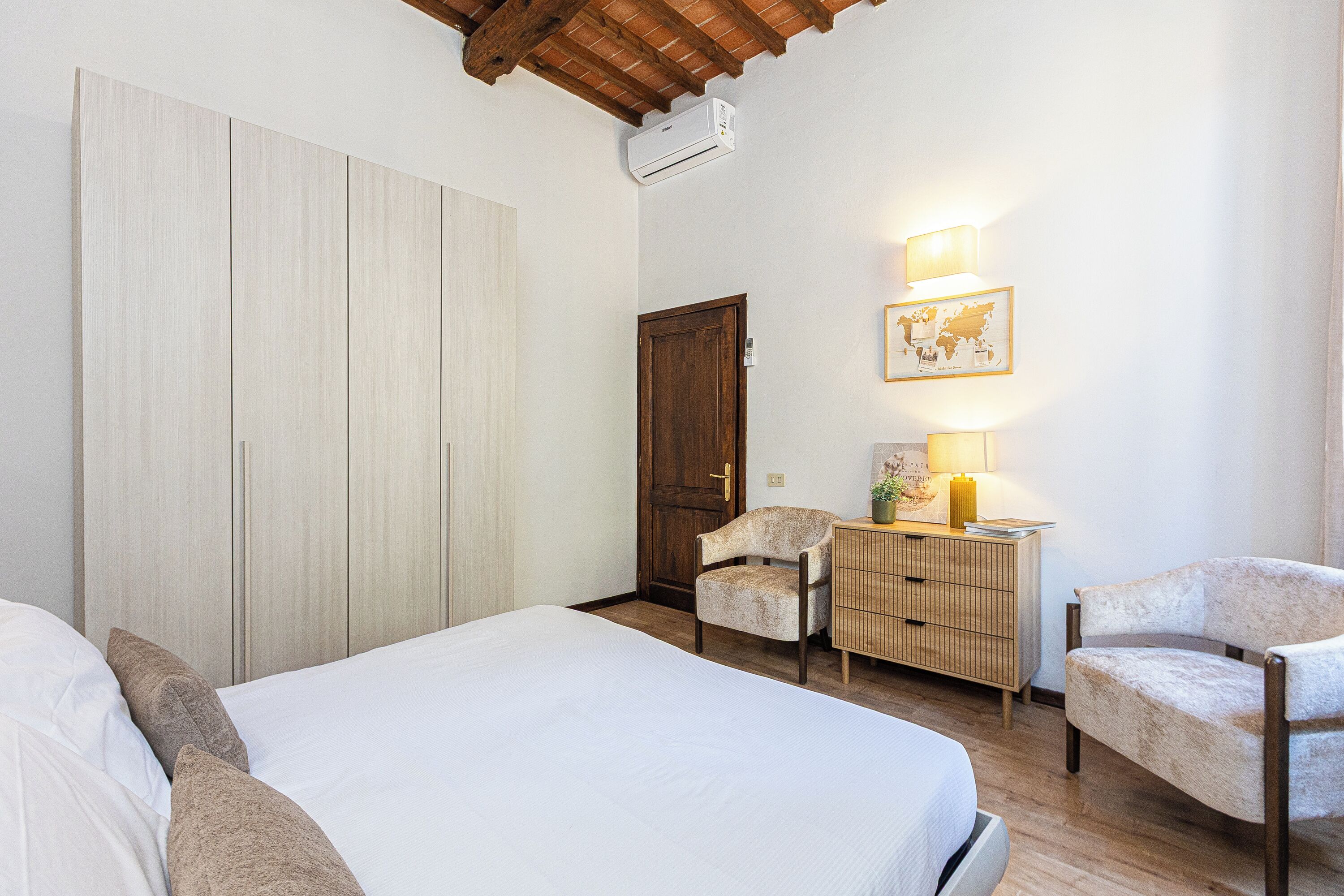 A bedroom with a bed, two chairs, a dresser, a wooden door, and a wall-mounted air conditioner.