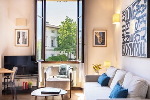 Uma sala de estar moderna com um sofá, uma poltrona, uma mesinha, uma televisão e uma janela ampla com vista para edifícios e árvores.