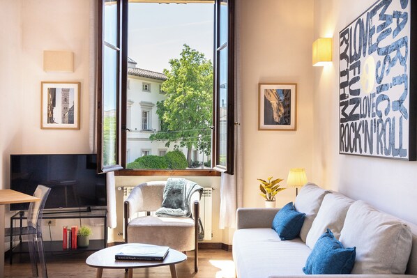 Uma sala de estar moderna com um sofá, uma poltrona, uma mesinha, uma televisão e uma janela ampla com vista para edifícios e árvores.