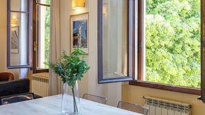 Une pièce comprenant une table, des chaises, un vase avec des plantes vertes, un tableau encadré et une fenêtre donnant sur des arbres.