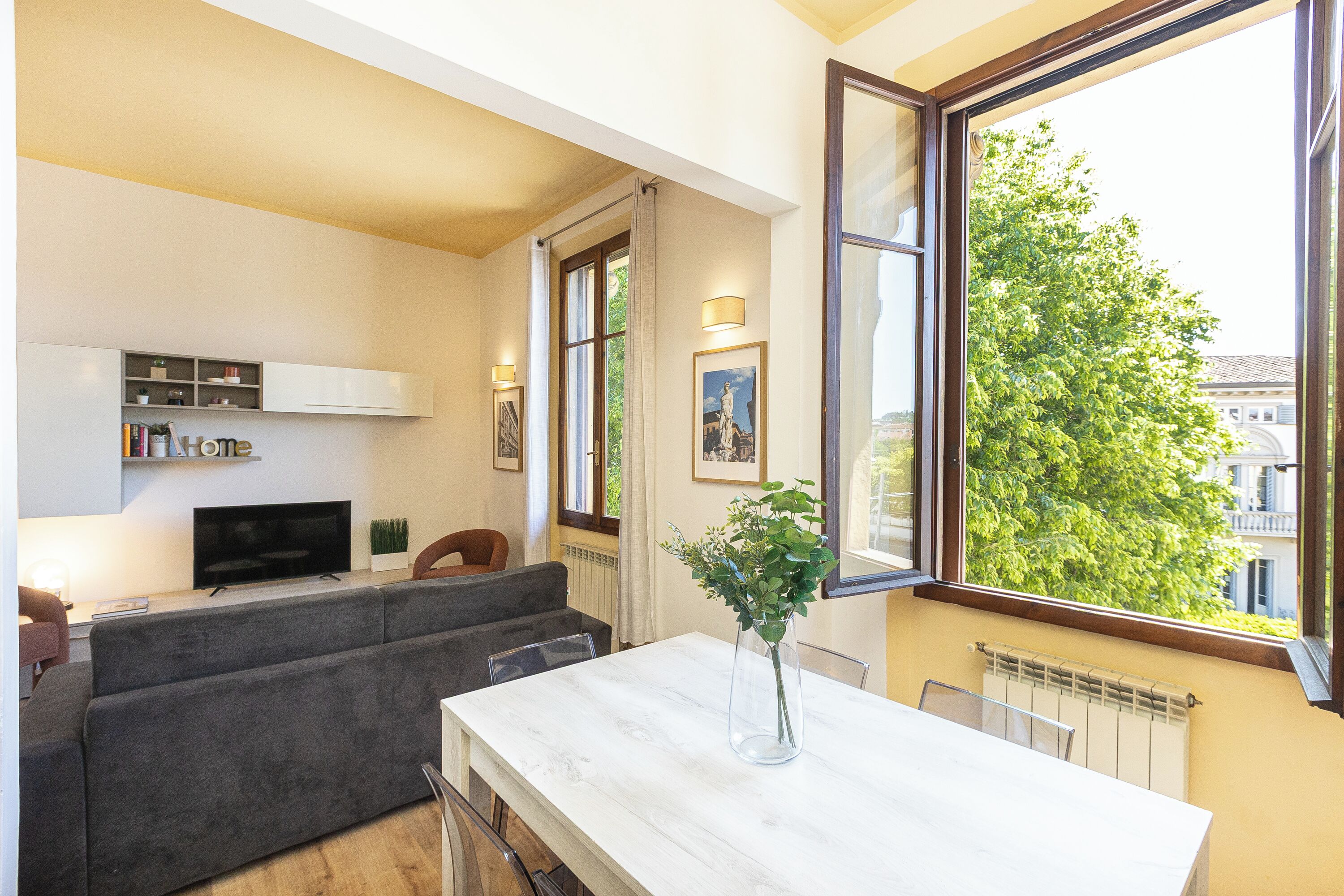 A modern living room with a sofa, a TV, a dining table, and a large window with a view of trees and buildings.