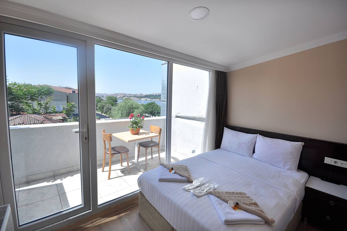 A hotel room with a large bed, a small table with chairs, and a view of the city through the balcony doors.