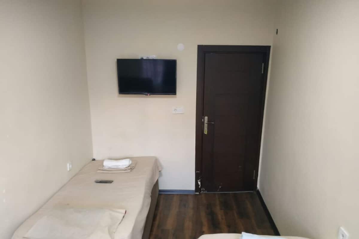 A small, single-bed room with a television mounted on the wall, a door, and a bedside table with a lamp.