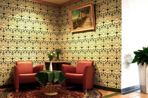A room with patterned wallpaper, a painting, two red armchairs, a small table with a green tablecloth, and a potted plant.