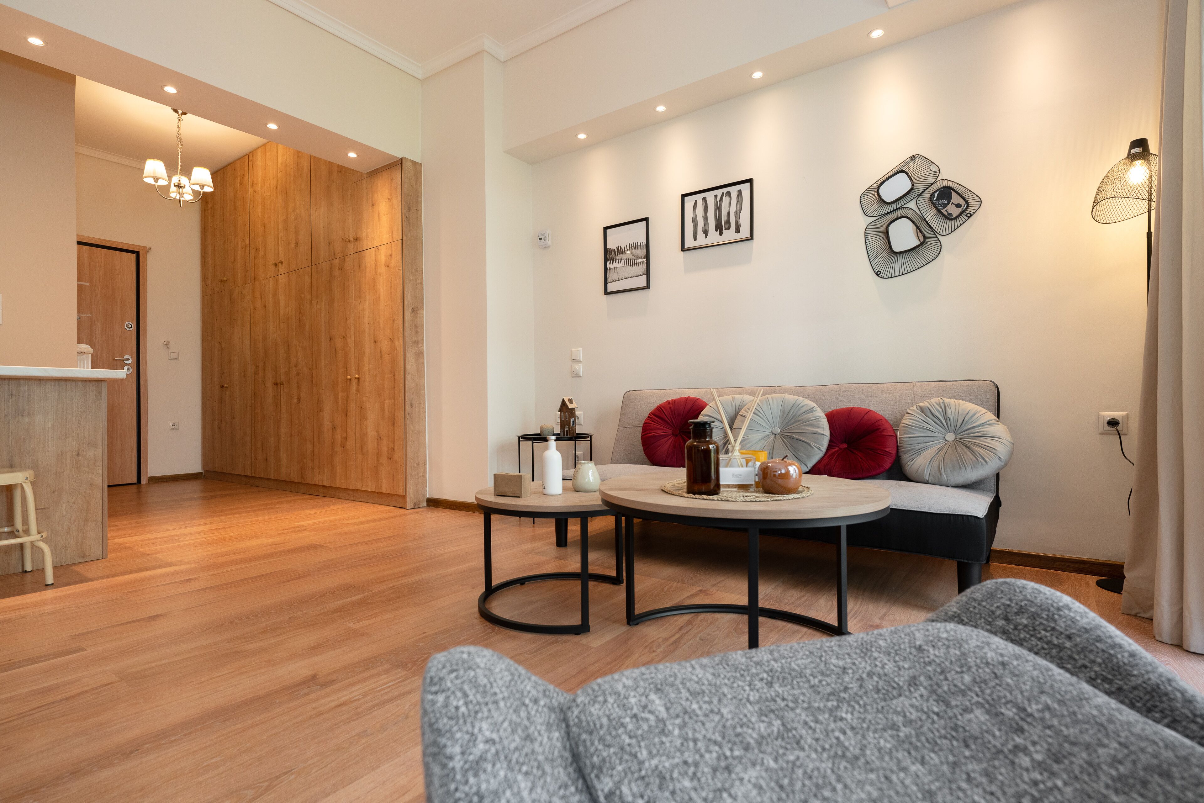A modern living room with a wooden floor, a sofa with red and grey cushions, a round coffee table, and a wall-mounted lamp.