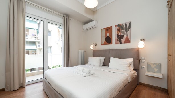 A neatly made bed with white linens and a pair of folded towels, a bedside table with a lamp, and a wall-mounted shelf with a small globe. - Contemporary Apartment made of Wood and Stone (Athens)