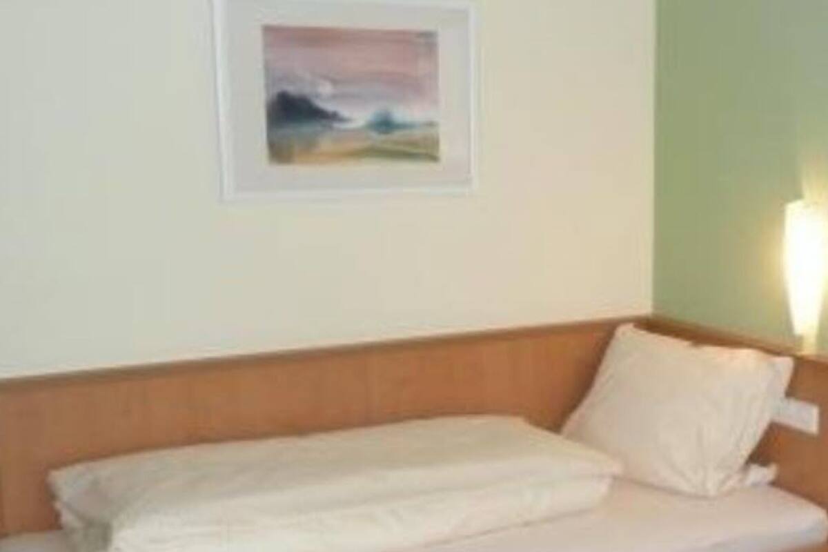 Una cama individual con ropa blanca, en una habitación con cabecera de madera, un cuadro enmarcado en la pared y una mesita de noche con lámpara.
